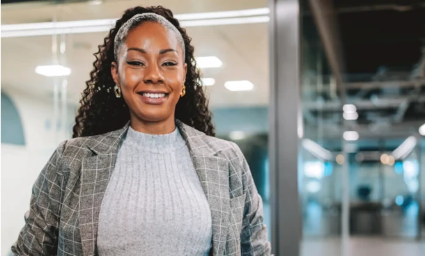 Smiling professional woman in a gray plaid blazer standing confidently in a modern office hallway.