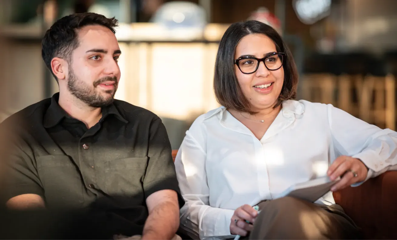 Two people sit together indoors, smiling and talking; one holds a notepad and pen, both look engaged.