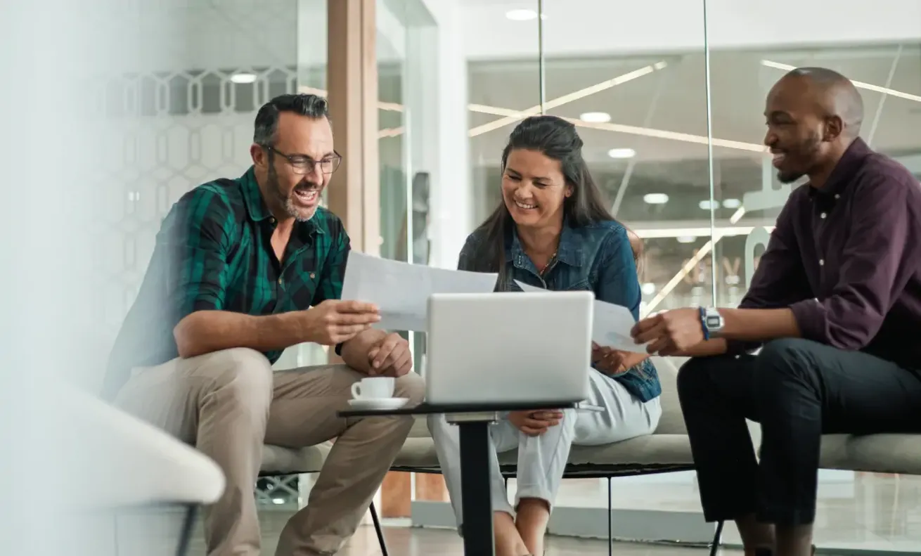Three smiling colleagues reviewing documents and a laptop during an informal meeting in a modern office lounge.